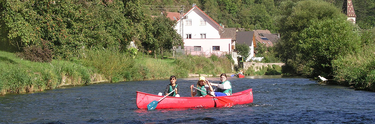 Im Vordergrund rotes Kanu quer, mit 3 Mädels; Blickrichtung auf den Fluss, im Hintergrund Weißes Haus am rechten Ufer, dovor zweites rotes Kanu auf dem oberen Neckar vor Fischingen